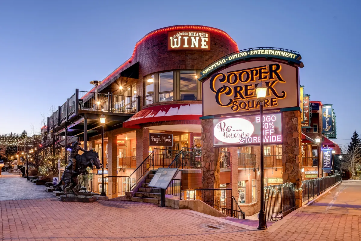 The bustling exterior of Cooper Creek Square, with shoppers and diners enjoying outdoor seating, visible signage for shops and wine bars, and a distinctive horse statue, all under lively, festive lighting.