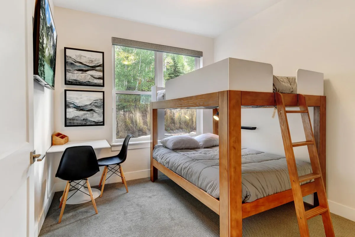 A comfortable bedroom designed for kids or teens, featuring a double-over-double bunk bed, a desk and chair for studying or quiet activities, playful artwork, and a window with a relaxing view. The room feels inviting, functional, and youth-friendly.