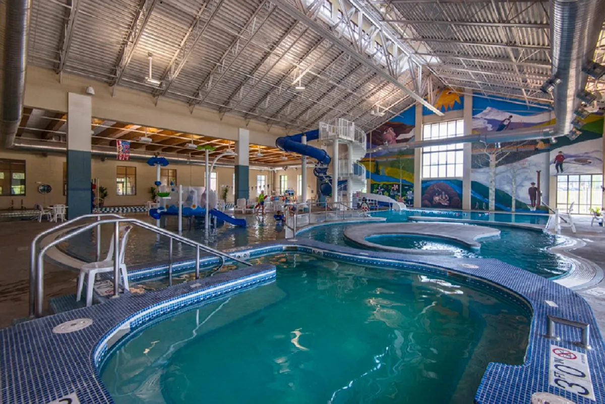 A lively, colorful indoor aquatic center featuring a large swimming pool, winding water slides, and playful mural artwork. Children and families are seen enjoying the water, radiating a joyful and energetic mood. The environment captures the fun, family-friendly appeal of local recreational amenities.