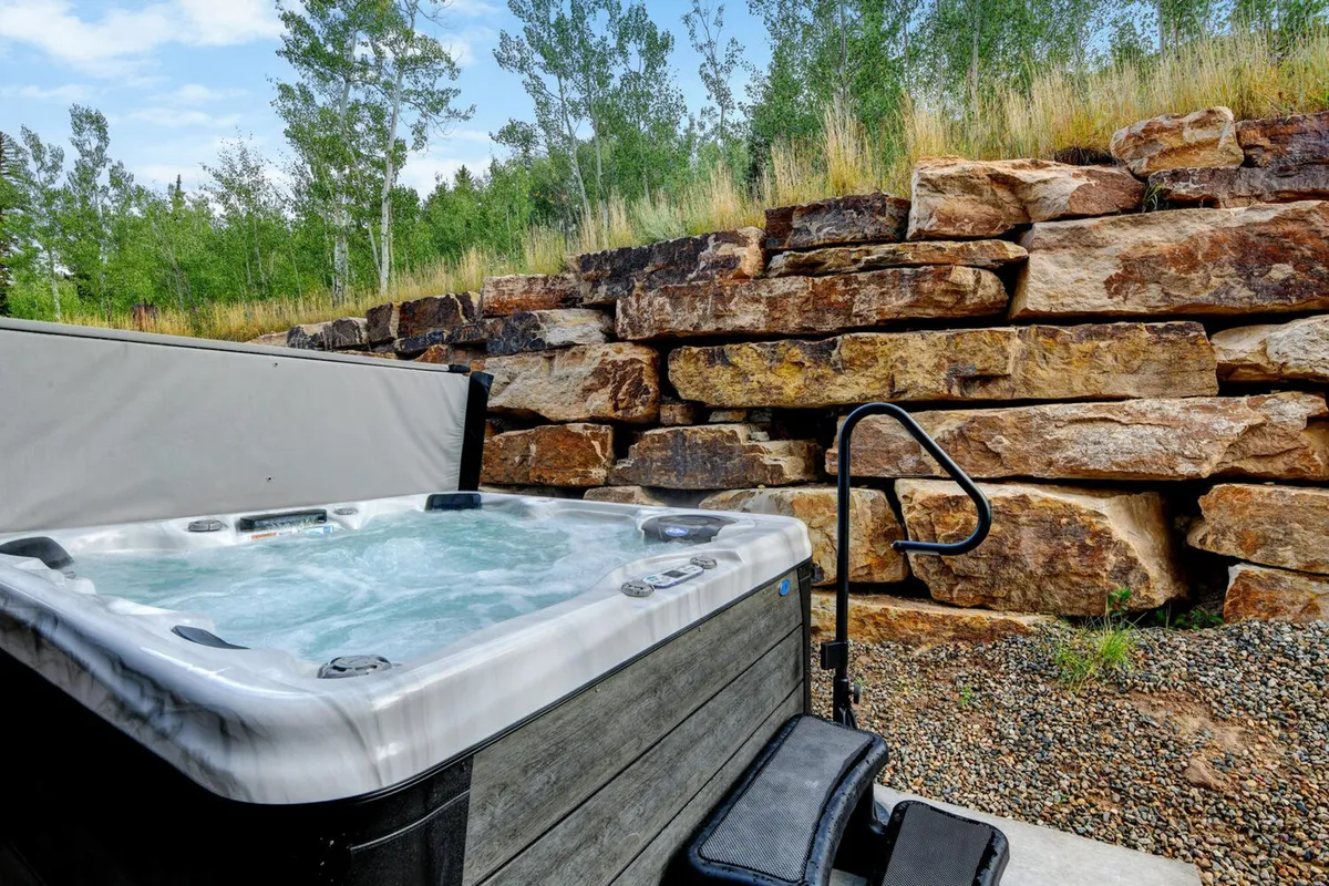 An inviting outdoor hot tub sits nestled against a rock wall, surrounded by lush greenery, creating a tranquil oasis. The scene suggests relaxation and privacy, with ample space for a group to unwind while soaking in nature and mountain ambiance.