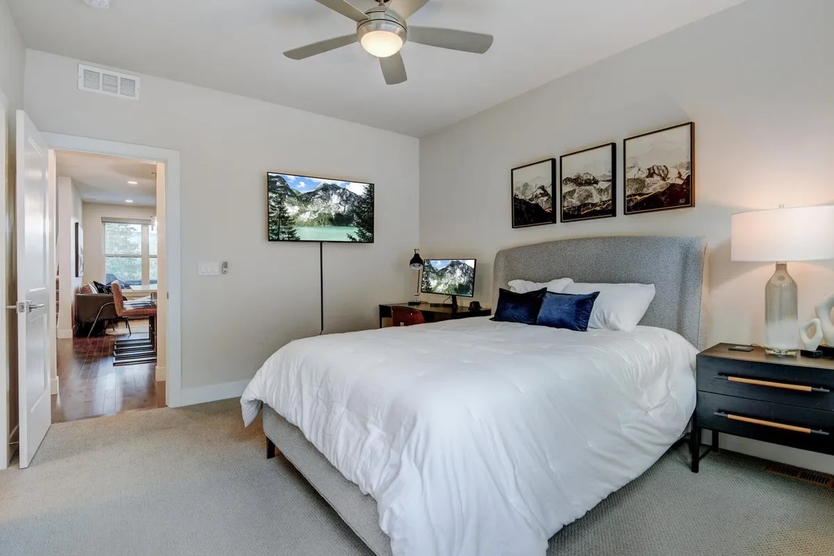 A cozy bedroom interior with a bed featuring white bedding, a mounted television, framed mountain landscape art, and a side table with a lamp. The mounted TV is a focal point, suggesting a comfortable spot for enjoying movies or sports events in style.