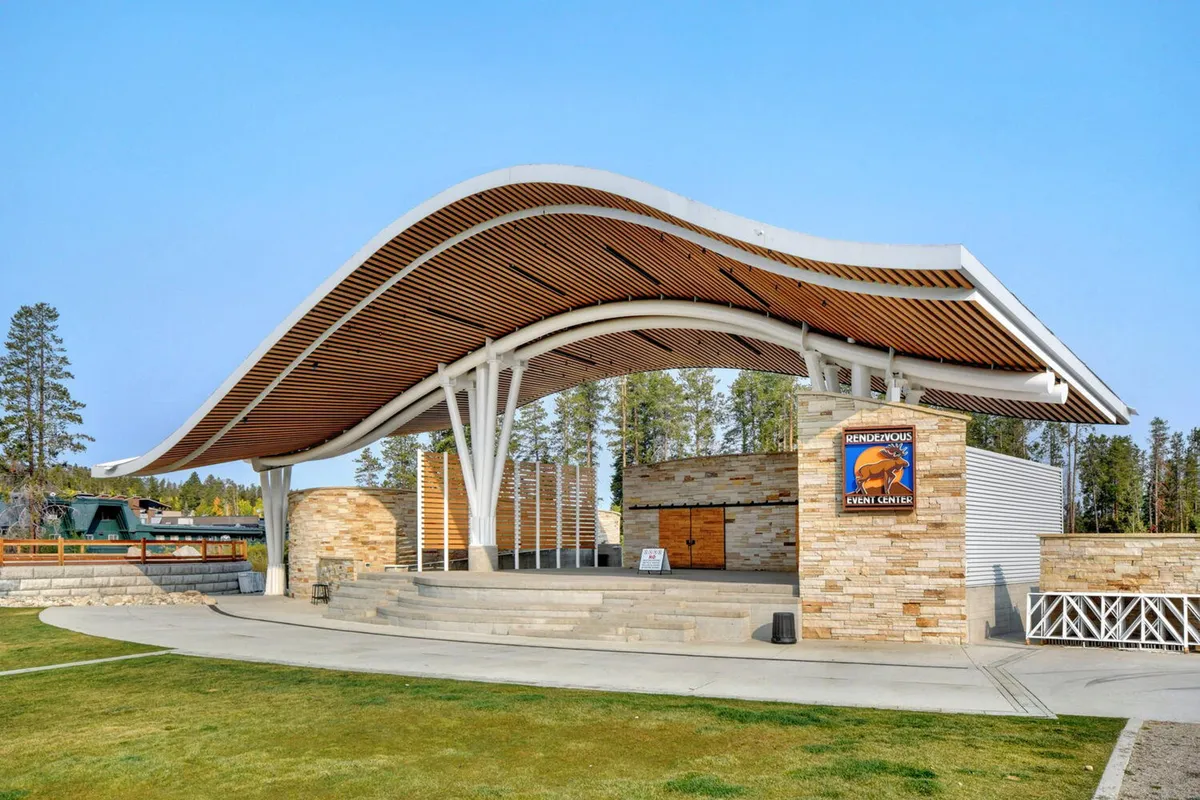 An exterior shot of an open-air amphitheater with a curved roof, stone walls, and a spacious stage area. The setting is designed for community events, concerts, and outdoor gatherings, with ample open space and a welcoming, communal vibe.