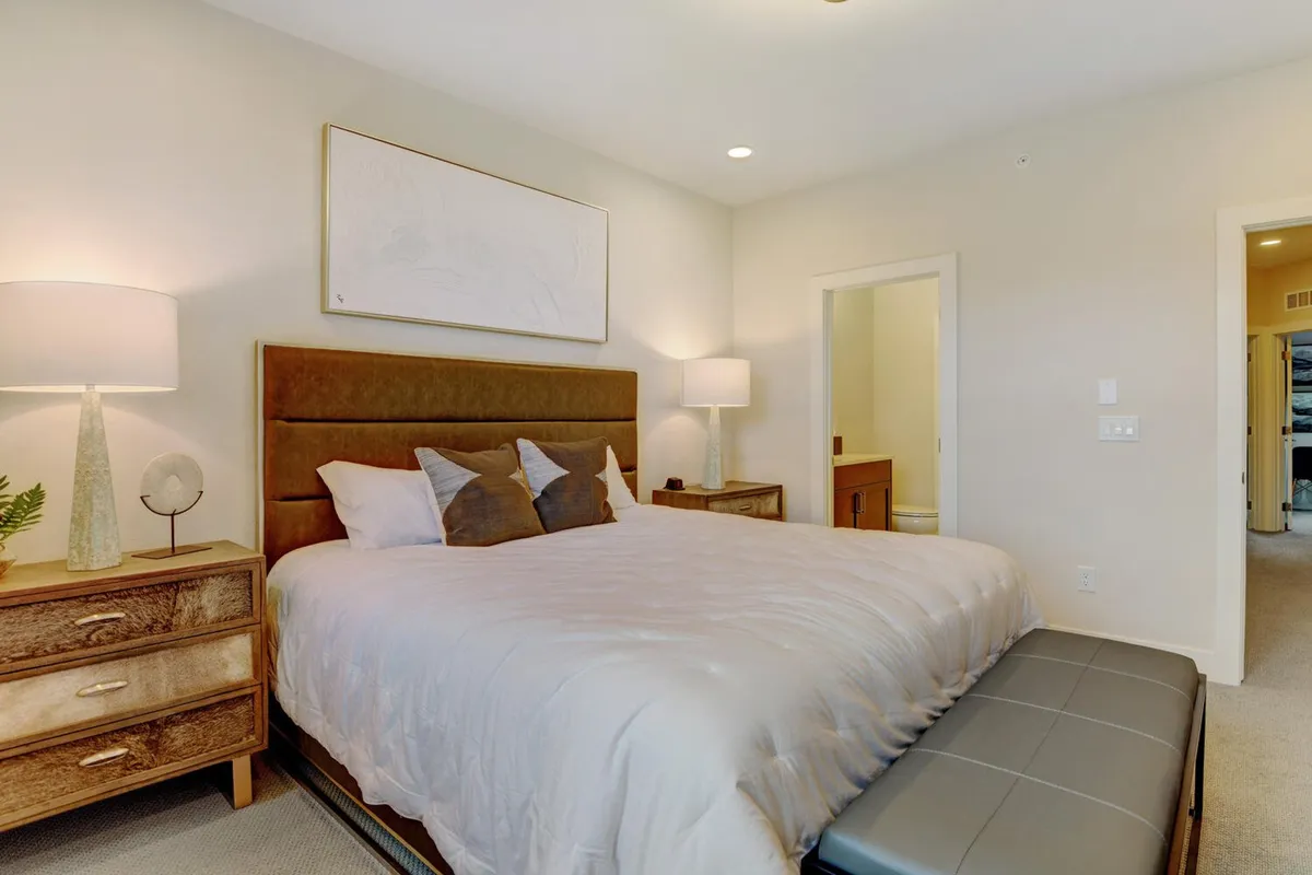 A tranquil bedroom featuring a plush bed topped with luxurious bedding, flanked by elegant nightstands with lamps, and highlighted by tasteful artwork above the headboard. Soft natural light filters through the window, creating a serene and restful ambiance.
