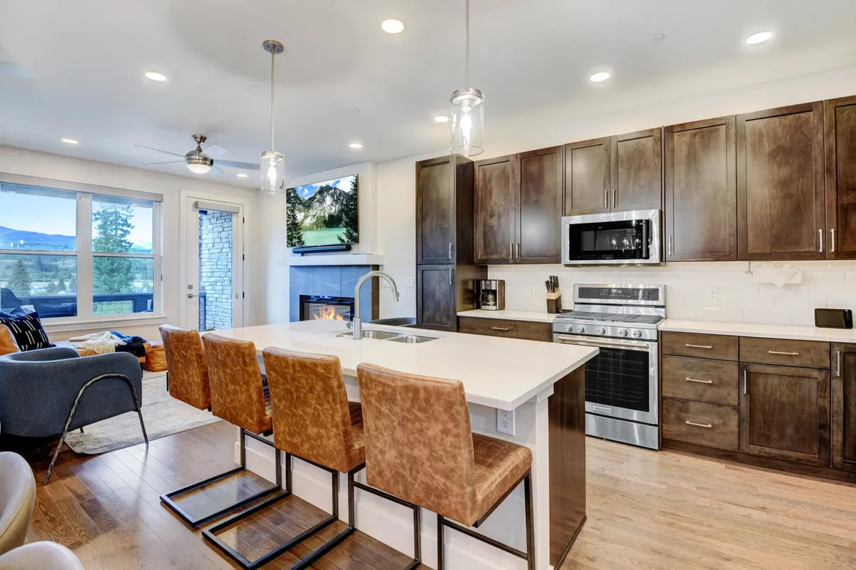 A warm, inviting kitchen and living space featuring a prominent kitchen island with bar stools, a cozy fireplace, a TV displaying a landscape scene, and large windows providing ample natural light and mountain views. The open layout fosters easy movement and interaction between spaces.