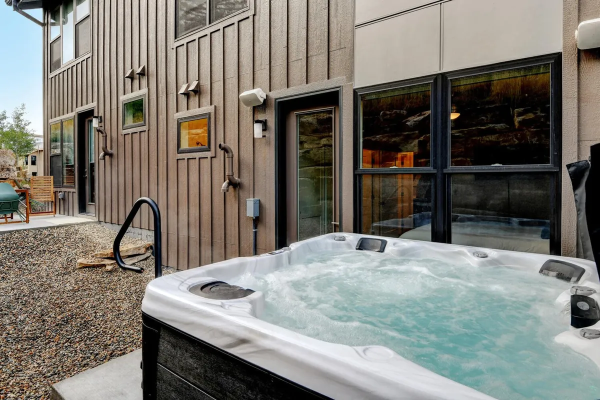 An outdoor space featuring a hot tub, modern siding, and ample windows. The scene is calm and inviting, hinting at both hot tub relaxation and casual socializing in the fresh mountain air.