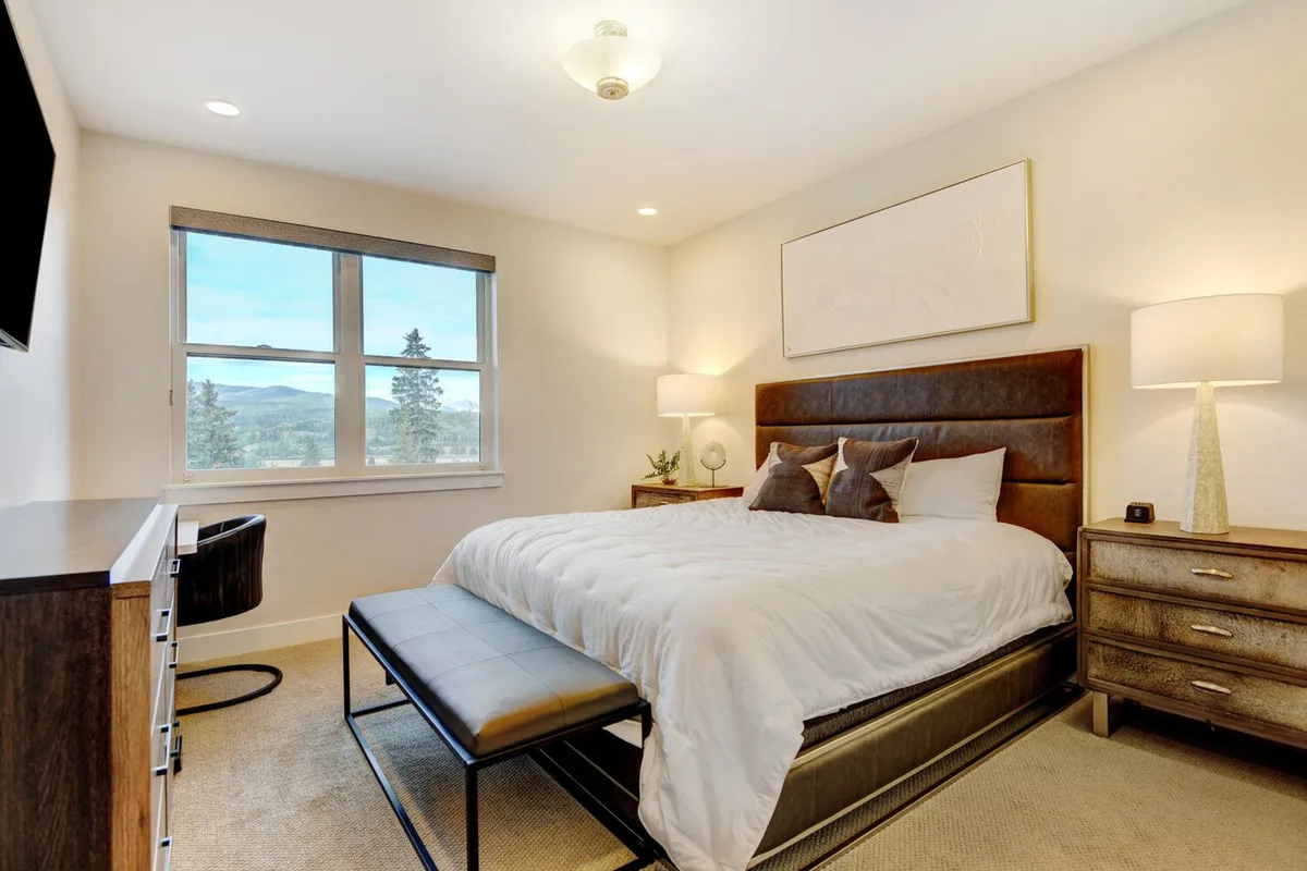 A cozy bedroom with a plush bed facing a large window that frames a beautiful scenic view. The space is complemented by a nightstand with a lamp and a dressing table, creating a calm and inviting space for relaxation and rejuvenation.