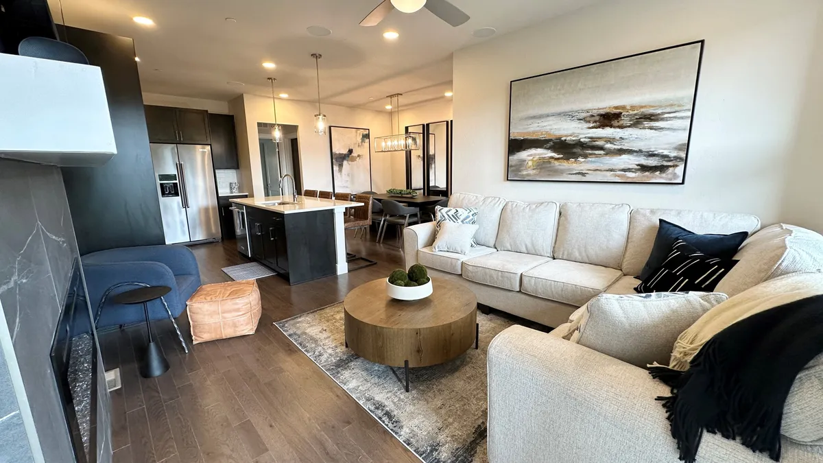 A spacious living room featuring a comfortable sectional sofa, a round wooden coffee table ideal for game nights, modern artwork on the wall, and a glimpse of the dining area in the background. The expansive windows (not explicitly mentioned, but implied by the mood and context) hint at natural light and mountain views, creating a cozy yet open atmosphere.
